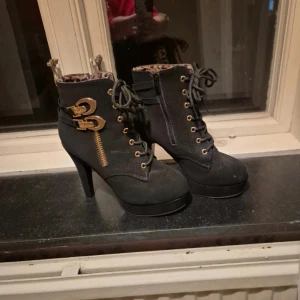 Svarta boots med klack - Säljer ett par svarta boots med hög klack från SHEIN. Skorna har snörning framtill, guldfärgade detaljer och dragkedja på sidan. Insidan är fodrad med leopardmönster.                   Storlek 37, 10cm hög klack (se bild för egen referens) Provade inomhus aldrig använda utomhus. Mycket fina i mocka imitation.  Nypris: 349kr  #klackskor #klackskor37 #skor37 #wilmabooksskor37