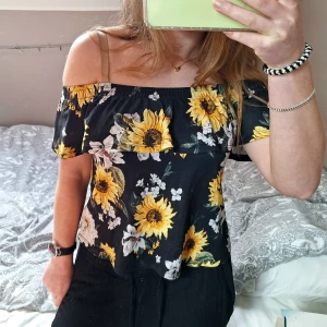 Svart offshoulder blus med solrosor - Superfin svart offshoulder blus från Gina Tricot som kan stylas på flera olika sätt. Blusen har stora gula solrosor och volang upptill. Perfekt för sommaren och våren. Jag använder den aldrig så den är i bra skick!🌻