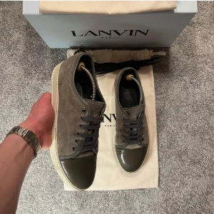Grå/ olivgröna sneakers från lanvin - Säljer ett par grå sneakers från Lanvin med ovandel i mocka och lackad tå i mörkgrönt. Skorna har snörning och vit platt sula. Klassisk och stilren design med exklusiv känsla. Passar både 41 42 men bäst på 42, hör av er vid minsta fundering! 😁bara skorna medföljer ej dust bag eller box!!