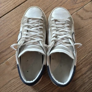 Vita sneakers från Philippe Model - Säljer ett par vita Philippe Model sneakers med svarta detaljer på hälen och grå mockadetaljer vid tån. Skorna har snörning och en platt sula. Klassisk och stilren design med rund tå och diskret logga på sidan