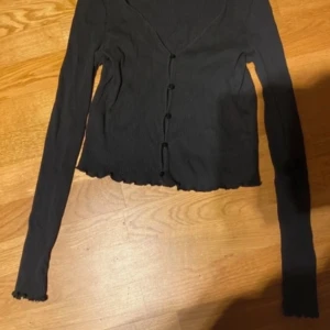 Svart ribbad kofta från H&M - Svart ribbad kofta från Vero Moda med v-ringning och knappar framtill. Modellen är croppad med långa ärmar och vågig kant nertill och vid ärmslut. Perfekt att styla till jeans eller kjol.