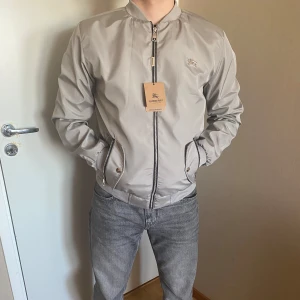 Burberry Windbreaker - Snygg ljusgrå windbreaker från Burberry med dragkedja framtill och ribbade muddar. Jackan har två fickor med knapp och diskret broderad logga på bröstet. Perfekt för dig som vill ha en stilren och klassisk look nu inför de härliga vädret 😊