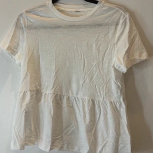 Vit t-shirt med volang - Säljer en vit t-shirt med rund hals och korta ärmar. T-shirten har en volangdetalj nedtill som ger en avslappnad och trendig look. Perfekt att matcha med jeans eller kjol.