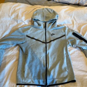 Grå hoodiejacka från Nike - Säljer en grå hoodiejacka från Nike med svarta detaljer och dragkedja framtill. Jackan har huva, lång ärm och en praktisk ficka med dragkedja på ena ärmen. Perfekt för en sportig och avslappnad stil.