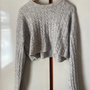 Grå croppad stickad tröja från Bershka - Säljer en grå croppad stickad tröja från Bershka med kabelstickat mönster. Tröjan har långa ärmar och rund halsringning, perfekt för lager-på-lager-stil. Endast använd 2 gånger