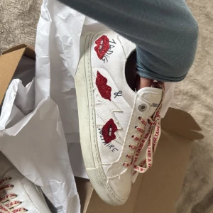 Vita sneakers från Zadig & Voltaire med röda detaljer - Säljer ett par vita sneakers från Zadig & Voltaire med röda glittriga läppar på sidan och röda snören med logga. Skorna har rund tå och platt sula, samt coola broderade detaljer. 😍😍Knappt använda tvättar av dom innan jag skickar så dom blir ännu renare! Fick dom i present men dom är för stora tyvärr!