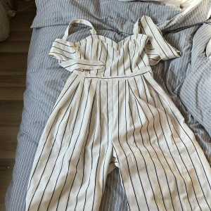 Vit randig axelbandslös klänning med knyt - Säljer en vit klänning med svarta vertikala ränder och axelbandslös design. Klänningen har en söt knytdetalj baktill och markerad midja. Perfekt för sommarens tillfällen!