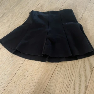 Svart kortkjol med utsvängd form - Säljer en svart kortkjol med utsvängd och voluminös siluett. Kjolen har en markerad midja och är sydd i ett slätt, något tjockare material som ger stadga. 