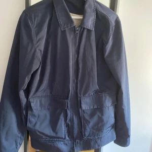 Windbreaker - En stilren mörkblå windbreaker med dragkedja framtill och två stora fickor. Klassisk krage och långärmad modell, perfekt för blåsiga dagar. Enkel och tidlös design som passar till många tillfällen.