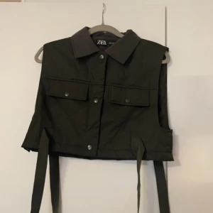 Mörkgrön väst från Zara med knappar - Säljer en mörkgrön väst från Zara med krage och knappar framtill. Västen har två fickor med lock och knappar på bröstet samt långa band i sidorna. Perfekt för att addera en cool touch till din outfit.