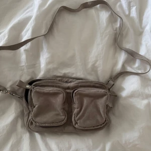 Beige axelväska med dubbla fickor - Säljer en beige axelväska i mocka med två framfickor med dragkedja och justerbar axelrem. Väskan har flera fack och detaljer i silverfärgad metall. Knappt använd!!!