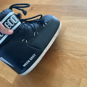 Svarta Moon Boots - Säljer ett par klassiska svarta Moon Boots med vit logga och snörning framtill. Skorna är i storlek 39-41. Skulle säga att de passar dem som har storlek 39. När jag köpte skona hade jag storlek 39 och de passade perfekt. Skorna är perfekta för kalla dagar. De är tillverkade i syntetmaterial och har en ikonisk design som sticker ut. De är i väldigt bra skick det ätnas är att knutorna på snörningen gått upp lite gran. Och att de har några de dejter på INSIDAN av skon😊skorna är köpta för 2000kr. 