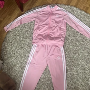 Rosa Adidas träningsbyxor med vita ränder och en matchande tröja till - Säljer ett par ljusrosa Adidas träningsbyxor och en ljusrosa tränings tröja med klassiska vita ränder längs sidorna och Adidas-logga på låret och bröstet . Byxorna har resår i midjan och är raka i modellen och tröjan är med drag kjedja . Perfekta för sport eller chill.