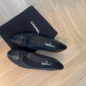Svarta ballerinaskor från Chanel - Säljer ett par klassiska svarta ballerinaskor från Chanel i skinn med rosett framtill och präglad logga på tån. Skorna har rund tå och platt sula, och kommer med originalpåse. Perfekta för en stilren look. De är i storlek 39,5 men jag är vanligtvis 39 och de sitter perfekt på mig! Endast använda en gång.
