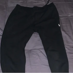 Svart hoodie från Polo Ralph Lauren - Svart hoodie från Polo Ralph Lauren med dragkedja framtill och klassisk logga på bröstet. Tröjan har huva med snörning och två fickor framtill. Perfekt för en avslappnad stil. Storlek m på koftan och S på byxorna, byxorna är stora i storleken 