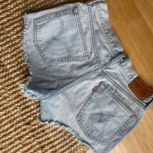 Ljusblå jeansshorts från Levi's - Säljer ett par ljusblå jeansshorts från Levi's med råa, fransiga kanter. Shortsen har låg/mid  midja och en avslappnad passform, perfekt för varma dagar. Ikonisk Levi's-lapp baktill och slitna detaljer framtill.