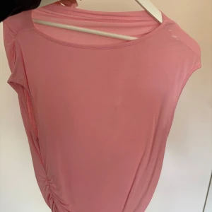 Rosa ärmlös topp med rynkdetalj - Säljer en ljusrosa ärmlös topp med rund halsringning och snygg rynkdetalj på ena sidan. Toppen är tillverkad i ett mjukt och stretchigt material som sitter bekvämt mot kroppen. Perfekt för varma dagar eller som ett lager under en jacka.