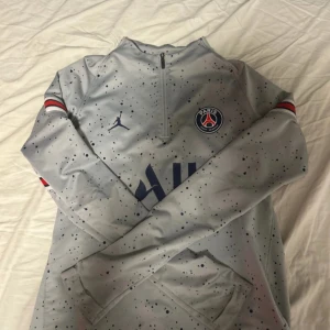 Grå träningströja från Paris Saint-Germain - Säljer en grå långärmad träningströja från PSG med svartprickigt mönster och halv dragkedja framtill. Tröjan har klubbens logga på bröstet och diskreta röda och blå detaljer vid ärmsluten. Perfekt för fotbollsträning.ÄKTA
