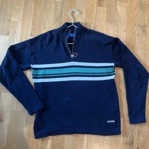 Marinblå tröja med dragkedja från Jack & Jones - Säljer en marinblå långärmad tröja från Jack & Jones med halv dragkedja och ringdetalj. Tröjan har blå, grå och gröna ränder över bröstet. Perfekt för en avslappnad stil och passar till många tillfällen.