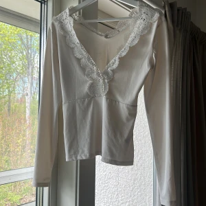 Vit långärmad topp med spetsdetaljer - Säljer en vit långärmad topp med v-ringning och fina spetsdetaljer längs halsringningen. Toppen har en feminin och elegant look med figurnära passform. Perfekt att styla till olika outfits.