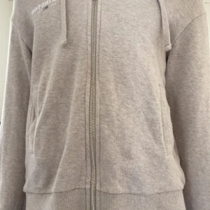 Beige hoodie med dragkedja från Lexington - Mysig beige hoodie från Lexington med dragkedja framtill och huva. Tröjan har två fickor på sidorna och ribbade muddar vid ärmslut och nederkant. Perfekt för en avslappnad stil.