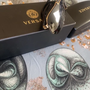Svarta solglasögon från Versace med gulddetaljer - Säljer ett par snygga svarta solglasögon från Versace med breda, mörka glas och exklusiva gulddetaljer upptill. Modellen har en futuristisk shield-form och ger en riktigt cool look. Perfekt accessoar för att sticka ut.