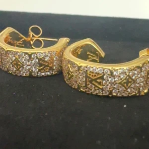 Guldiga örhängen från Lulu med stenar - Säljer ett par exklusiva guldiga örhängen från Lulu. Örhängena är halvbågsformade och dekorerade med gnistrande stenar samt klassiska LV-loggor och monogramdetaljer. Perfekta för dig som vill sticka ut med lyxig accessoar.