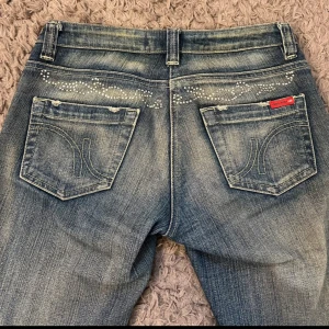 Blå jeansbyxor med glitter  - Säljer ett par klassiska blå jeans med snygga slitningar och dekorativa nitar på fickorna. Byxorna har raka ben och normal passform, samt tydliga sömmar och broderade bakfickor med logga. Storlek Small. Midjemått 35cm. Innerbensmått 78
