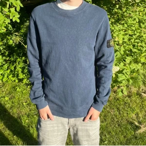 Blå sweatshirt från Stone Island - Säljer en blå sweatshirt från Stone Island med klassisk patch på vänster ärm. Tröjan har rund halsringning och långa ärmar. Perfekt för dig som gillar streetwear och vill ha något stilrent men ändå med tydlig logga. Storlek Small