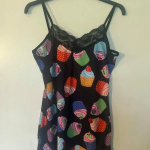 Linne M från n Insanity - Säljer en svart klänning med färgglatt cupcake-mönster och tunna axelband. Klänningen har spetsdetalj vid urringningen och är perfekt för dig som gillar lekfull stil. Materialet känns mjukt och lätt.