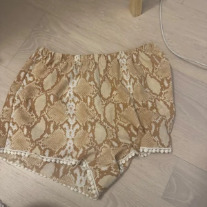 Beige shorts med ormmönster och dekorativ kant - Säljer ett par beige shorts med ormmönster och elastisk midja. Shortsen har en dekorativ kant med små bollar längst ner och är perfekta för varma dagar.