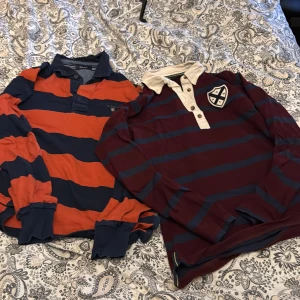Randiga rugbytröjor från Gant - Två klassiska rugbytröjor från Gant. Ena är orange och marinblå med breda ränder och krage, den andra är vinröd och marinblå med emblem på bröstet och vit krage. Båda har långa ärmar och knappar vid halsen. Perfekt för en sportig och avslappnad stil. 