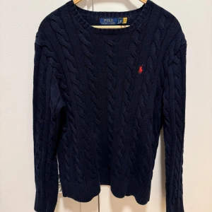 Mörkblå stickad tröja från Polo Ralph Lauren - Kabelstickad mörkblå tröja från Polo Ralph Lauren med rund halsringning och röd broderad logga på bröstet. Klassisk modell med långa ärmar och ribbade muddar. Nästan aldrig använd.  Perfekt för en stilren look. Ny pris 2 500 kronor. 
