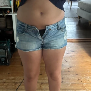 Blå jeansshorts med slitningar - Snygga blå jeansshorts med slitna detaljer och fransig kant nertill. Klassisk femficksmodell med låg midja och knappgylf. Perfekta för varma dagar och en avslappnad stil.