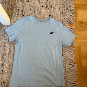 Ljusblå Nike t-shirt i bomull - Snygg ljusblå t-shirt från Nike med klassisk svart logga på bröstet. Enkel och stilren design med rund hals och korta ärmar. Perfekt för dig som gillar sportiga och avslappnade vibes. Tillverkad i mjuk bomull för skön känsla hela dagen.