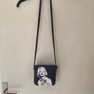 Svart axelväska med Marilyn Monroe-tryck - Cool svart axelväska med  axelband och ikoniskt Marilyn Monroe-tryck på framsidan. Väskan har vit kontrastsöm och en stilren, fyrkantig form. Perfekt för dig som vill sticka ut med en unik accessoar. 15*14 cm