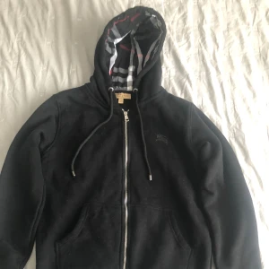 Svart burberry hoodie - Svart hoodie från Burberry med klassiskt rutigt foder i huvan och dragkedja framtill. Tröjan har snörning i huvan, känguruficka och långa ärmar. Perfekt för dig som gillar stilrena och exklusiva plagg. Pris kan diskuteras 