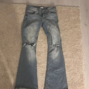 Ljusblå bootcut jeans med slitningar - Snygga ljusblå jeans med bootcut passform och slitna hål över knäna. Jeansen har klassisk femficksdesign, normal midja och är tillverkade i mjukt denimtyg. Perfekta för en avslappnad och trendig look.
