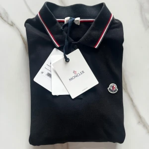 Moncler Piké Maglia svart - Svart långärmad pikétröja från Moncler med klassisk krage och knäppning. Tröjan har Moncler-logga på bröstet och röda/vita detaljer på krage och ärmslut. Tillverkad i bomull för en skön och stilren känsla. Perfekt för dig som gillar exklusiva kläder. Storlek XL men passar L. Obs lagad reva som syns på sista bilden. Skriv för fler bilder.