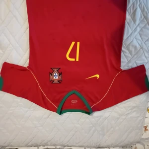 Portugal C. Ronaldo 17 Nike fotbollströja - Säljer en klassisk Portugal fotbollströja från Nike med C. Ronaldo och nummer 17 på ryggen. Tröjan är röd med gula detaljer, gröna muddar och krage samt Portugals landslagsemblem på bröstet. Perfekt för dig som älskar fotboll och Ronaldo.