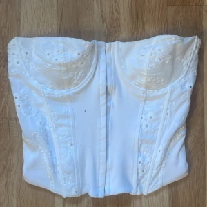 Vit korsett-topp från Zara - Supersnygg vit korsett-topp från Zara med broderade blommor och håldetaljer. Toppen är axelbandslös och storlek S.🙌🏻Passar dock Xs-M oxå skulle jag säga. Materialet är stretchigt och korsetten har inga tecken på användning.❤️ Tryck gärna köp knappen!☺️
