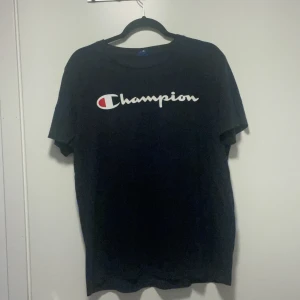 Svart Champion t-shirt med logga - Svart t-shirt från Champion med klassisk vit och röd logga tryckt över bröstet. Enkel och stilren design med rund hals och korta ärmar. Perfekt för dig som gillar streetwear och sportiga vibes.