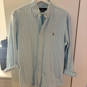 Blåvit randig skjorta Ralph Lauren M - Snygg blå skjorta med vita ränder från Ralph Lauren. Klassisk button-down krage, broderad logga på bröstet och slim fit passform. Långärmad och tillverkad i bomull, perfekt för en stilren look. 