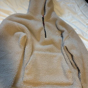 Beige fleece hoodie från Hugo - Säljer en beige fleece hoodie från Hugo med broderad logga på bröstet. Tröjan har huva, lång ärm, magficka och en svart half zip-dragkedja framtill. Perfekt för kalla dagar när du vill vara både varm och snygg.