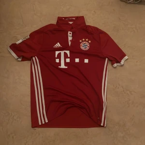 Bayern München Robben 10 Adidas tröja - Röd Bayern München matchtröja med vita detaljer, Adidas-logga och klubbmärke på bröstet. Nummer 10 och Robben tryckt på ryggen. Klassisk krage med knappar och vita ränder längs sidorna. Perfekt för dig som älskar fotboll och vill hylla Robben.