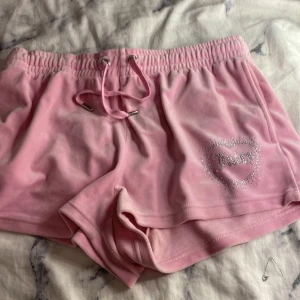 Rosa Juicy Couture velourshorts - Supergulliga rosa velourshorts från Juicy Couture med glittrande strasstenar som bildar texten 'Juicy' både bak och fram i ett hjärta. Shortsen har snörning i midjan och är perfekta för en chill vibe. Mjukt material och klassisk Juicy-stil.