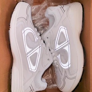 Vita Dior B30 sneakers med reflexdetaljer - Snygga vita Dior B30 sneakers med reflekterande detaljer och tydlig Dior-logga på sidan. Skorna har en chunky sula, meshpaneler och partier i syntet och skinn. Perfekta för dig som vill sticka ut med en exklusiv och modern look.