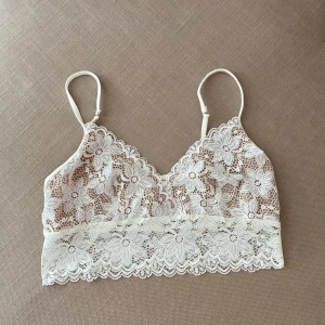 Vit spetsig linnetopp med blommönster - Superfin vit linnetopp i spets med stora blommor och tunna axelband. Toppen har v-ringning och är ärmlös, med en romantisk och feminin känsla. Perfekt att styla med jeans eller kjol för en trendig look.