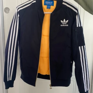 Svart bomberjacka från Adidas - Svart bomberjacka från Adidas med klassiska vita ränder längs ärmarna och broderad logga på bröstet. Jackan har dragkedja, ribbade muddar och en ficka med dragkedja på ena ärmen. Insidan är fodrad i gult för en extra cool kontrast.