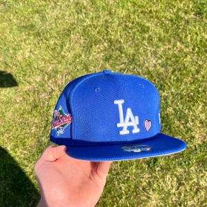 LA Dodgers keps New Era  - Snygg blå LA Dodgers keps från New Era 59FIFTY med broderad LA-logga framtill, hjärtdetalj och 1988 World Series patch på sidan. Baksidan har texten 'Love & Hustle = Game' och MLB-logga. Klassisk passform. Köparen betalar frakt